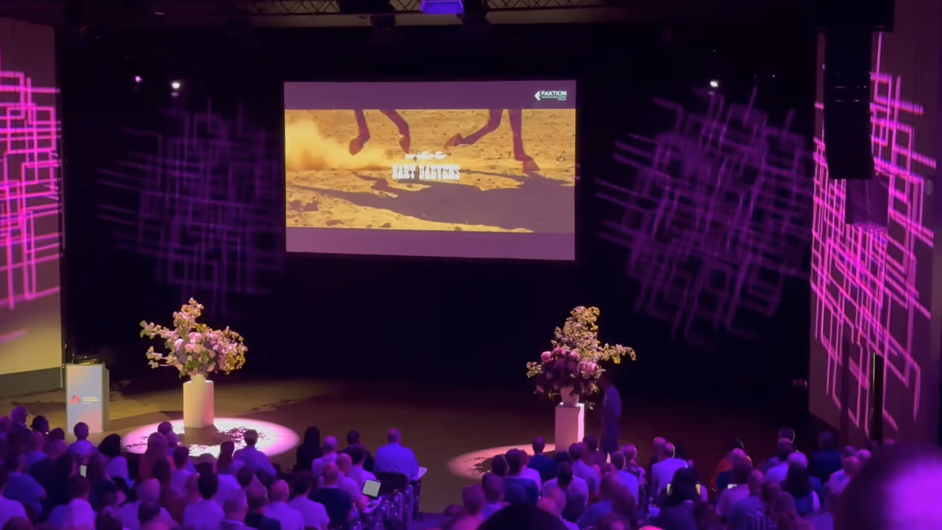 Audience seated in a dark theater watching a video projection of horse legs running on sand with purple geometric light patterns on side walls.