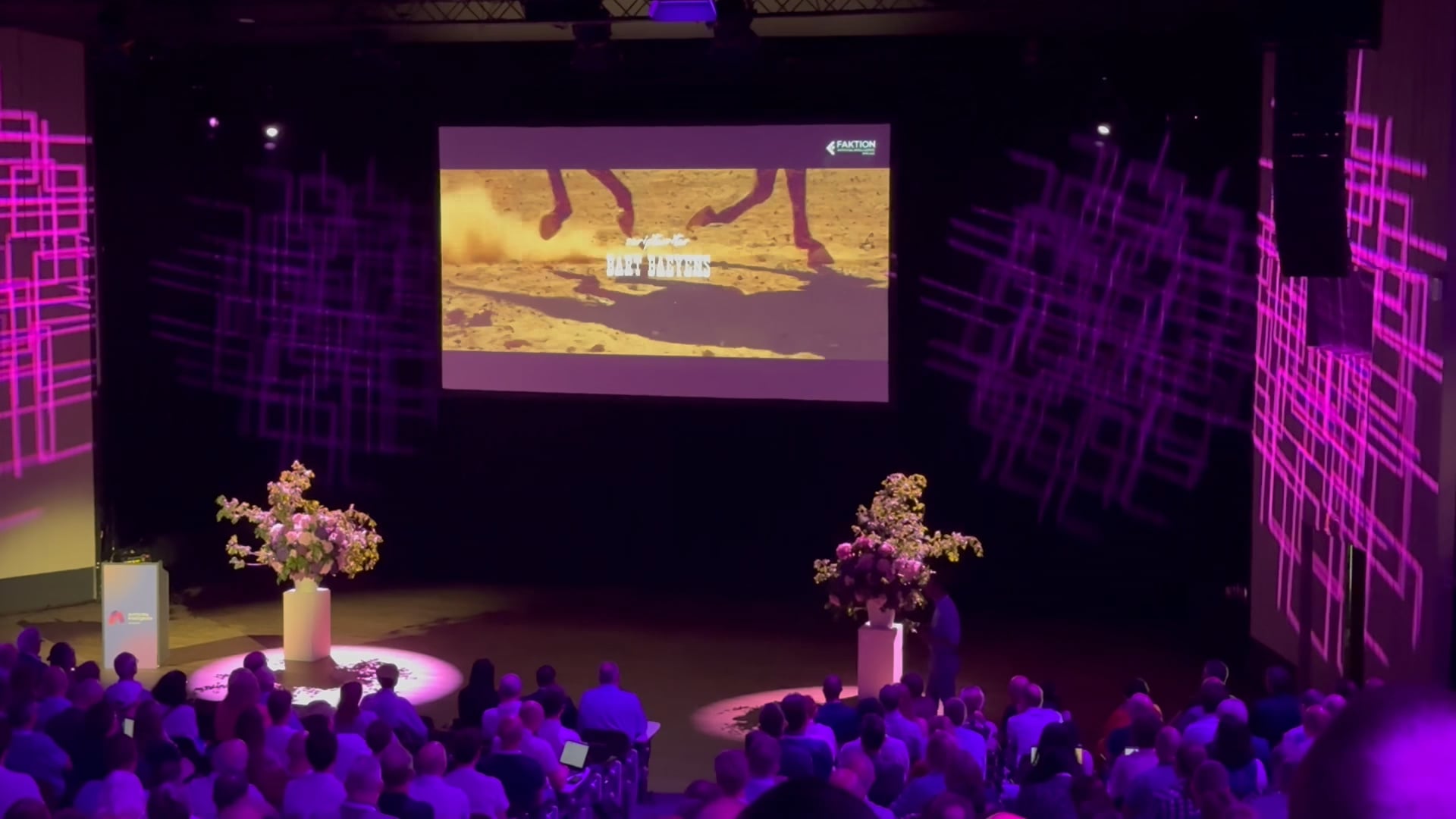 Audience seated in a dark theater watching a video projection of horse legs running on sand with purple geometric light patterns on side walls.