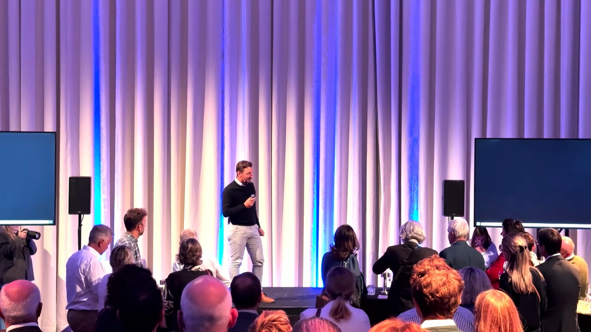 Man in black sweater and light pants speaking on stage to an audience standing in front of white curtains with blue lighting.