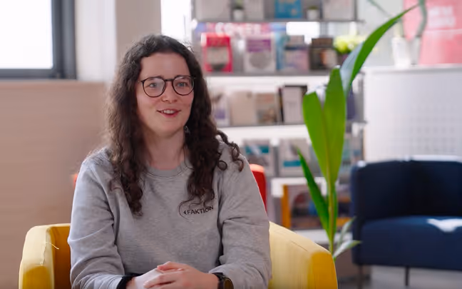 Woman with glasses and curly hair sitting on a yellow chair in a modern office setting.