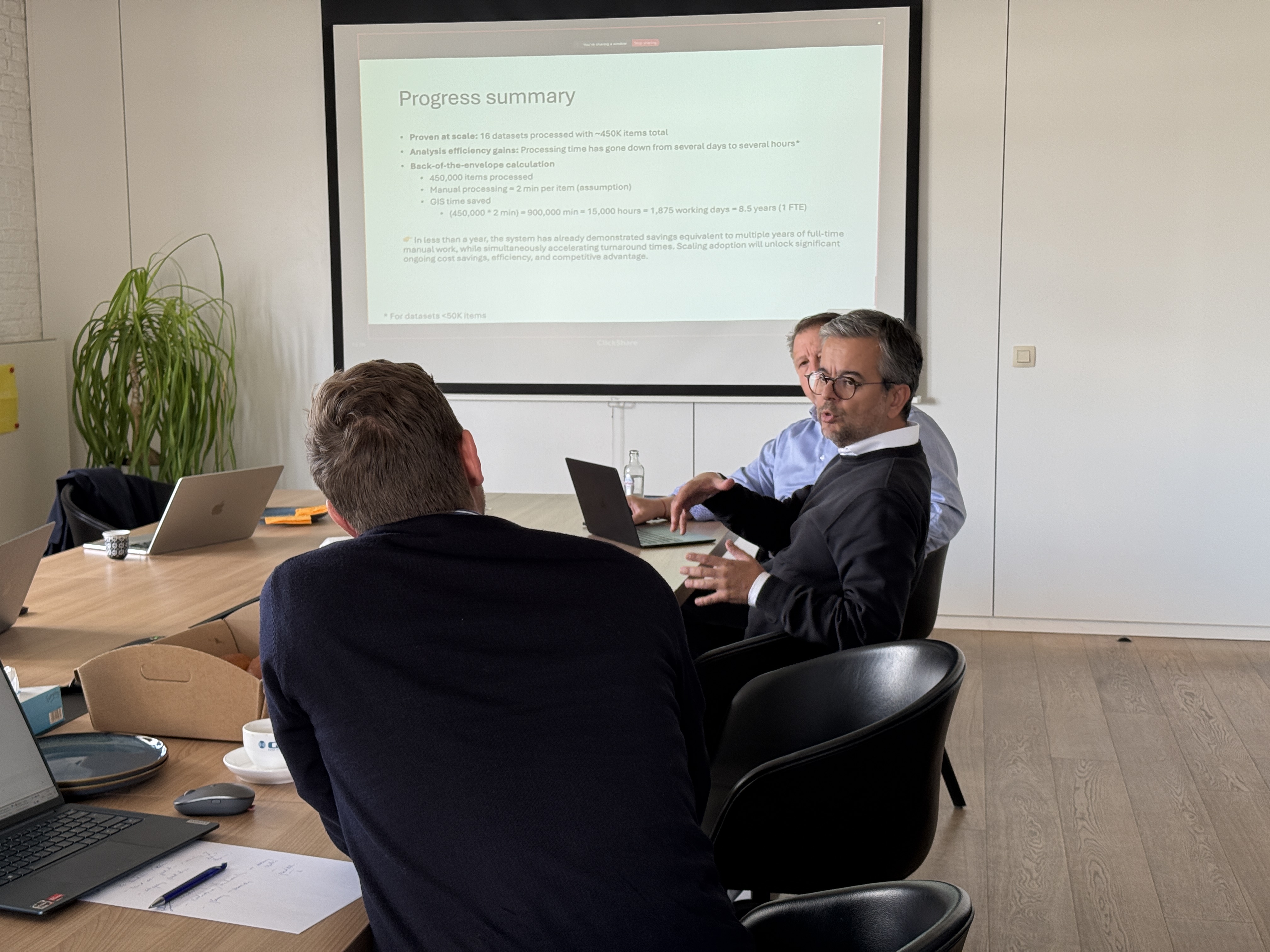 Three men in a meeting room discussing a presentation projected on a screen titled Progress summary.