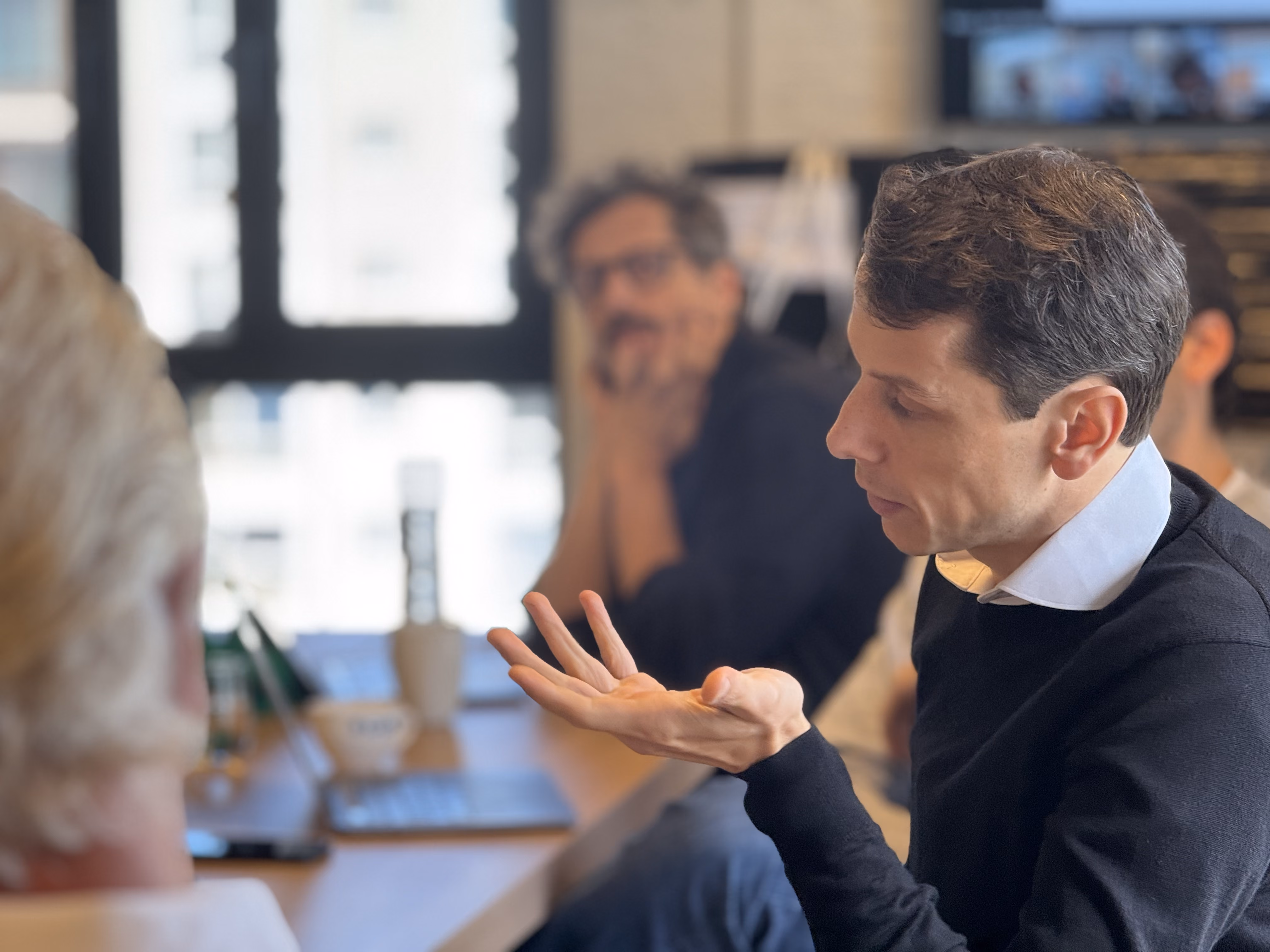 Man in black sweater speaking and gesturing with his hand in a meeting room with others.