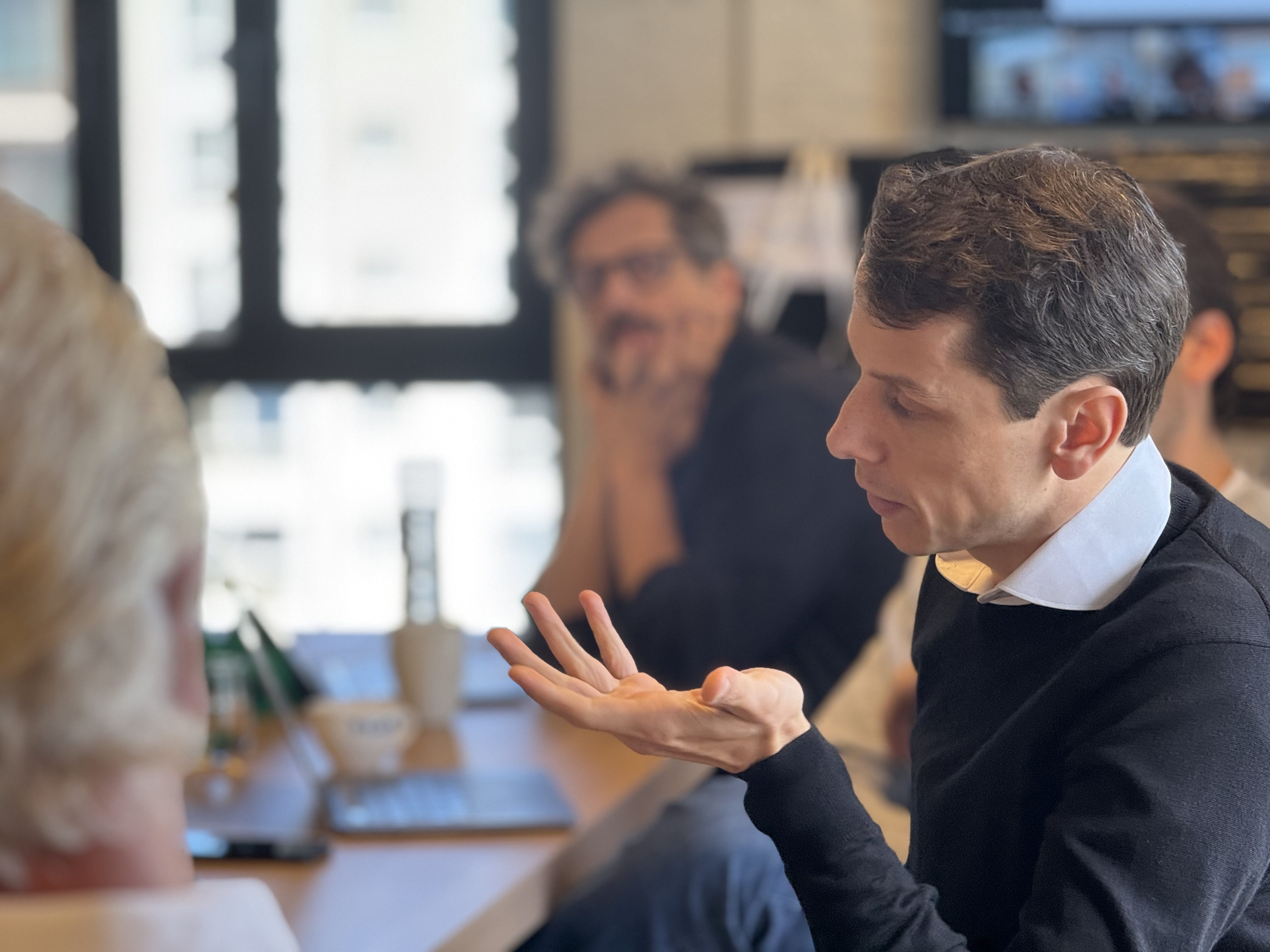 Man in black sweater speaking and gesturing with his hand in a meeting room with others.