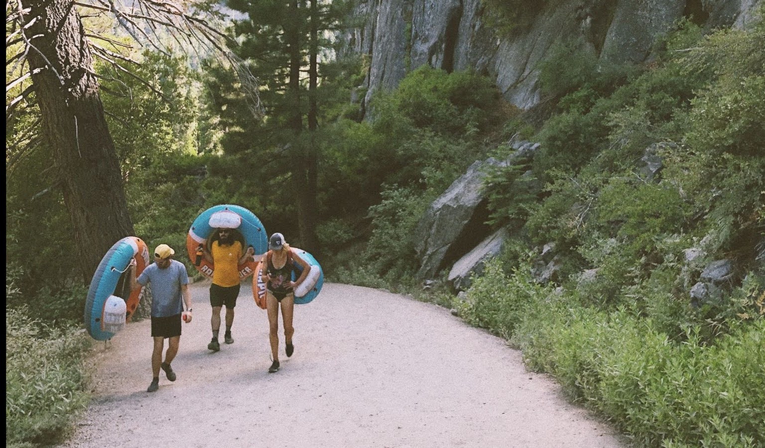 Three people carrying inflatable tubes along a gravel path beside a rocky cliff and pine trees
