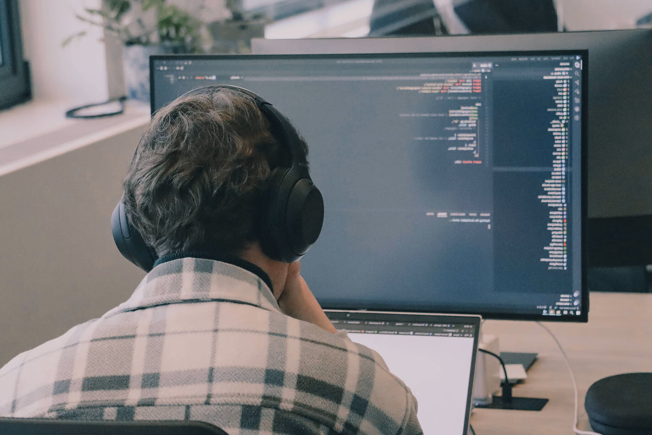 Persoon met koptelefoon kijkt naar computerschermen met programmeercode op kantoor.