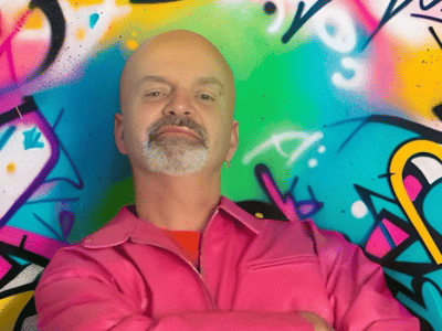 Xander in a pink leather jacket stood in front of a bright colour graffiti wall.