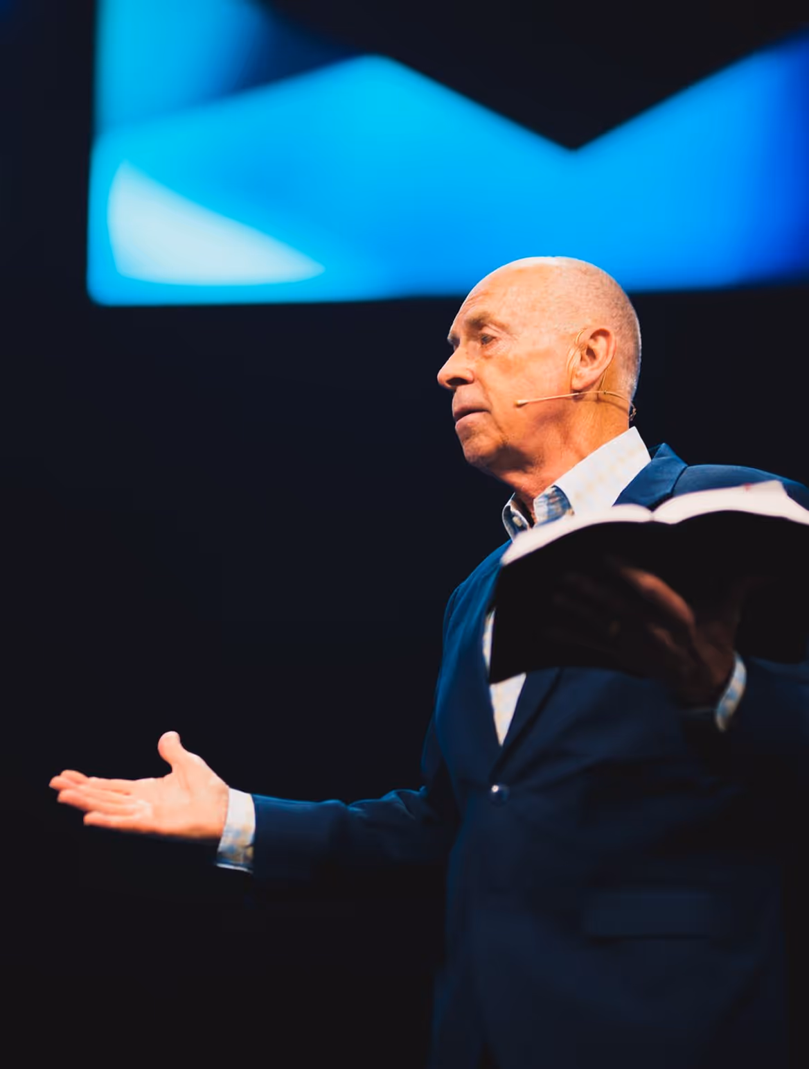 Older speaker on stage with blue background gesturing during presentation
