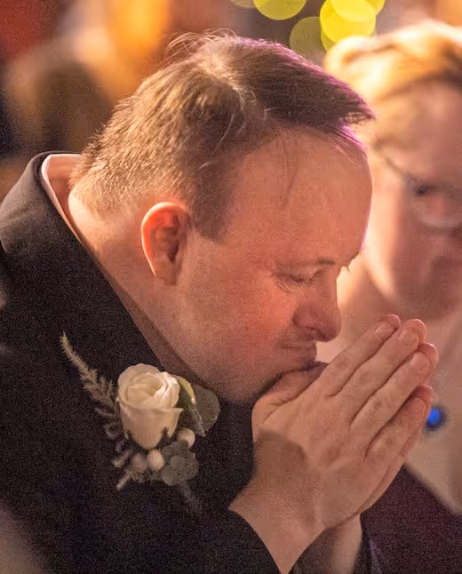 Person in formal attire praying with white rose boutonnière at emotional event