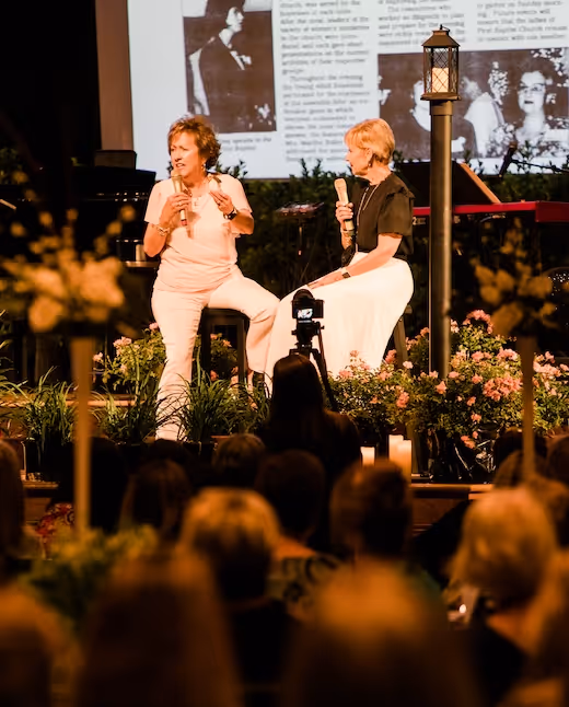 Two women speaking on stage surrounded by flowers and audience