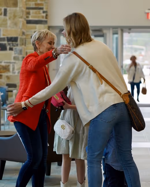 Two women warmly hugging in a bright, stone-walled interior space