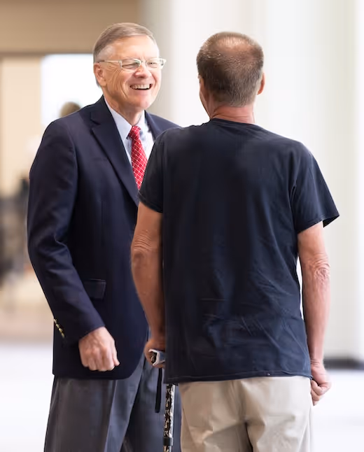 Two men talking, one in a suit smiling, the other wearing a black t-shirt