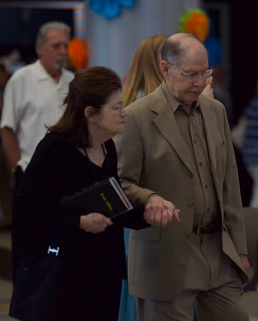 Older man in suit being guided by woman in black at event with balloons