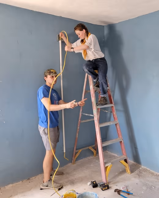 Two people working on electrical wiring inside a blue-painted room