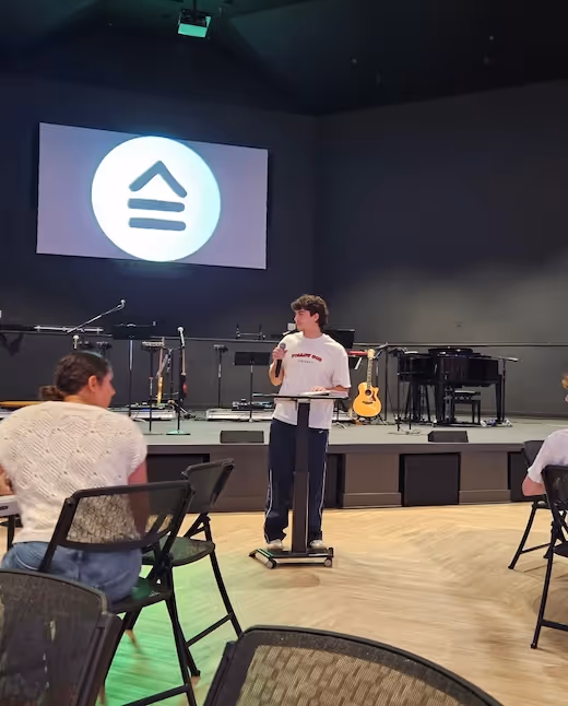 Person speaking at podium with logo screen and musical instruments behind