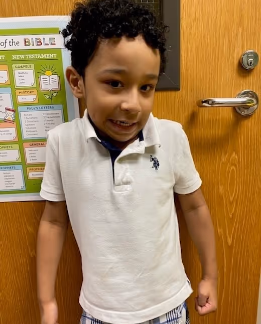 Smiling child in white polo shirt standing in front of Bible study poster