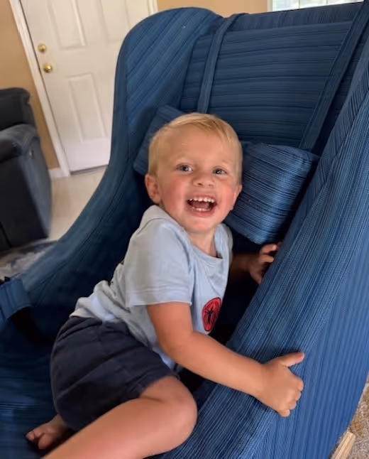 Smiling toddler hugging blue chair, wearing white shirt and blue shorts
