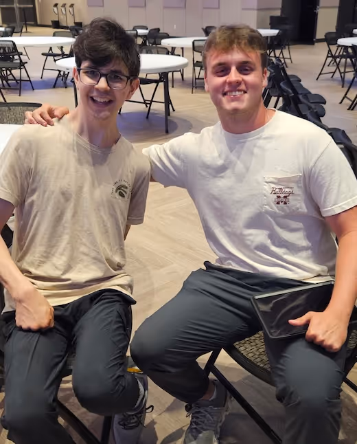 Two young men smiling and sitting together in a room with tables