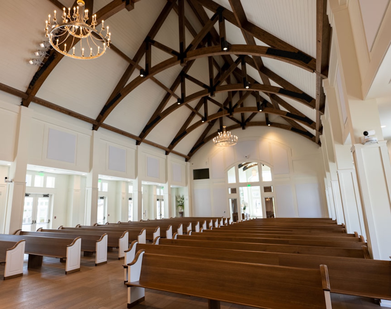 Interior of a spacious chapel with wooden pews, high white ceiling with visible wooden beams, and chandeliers.