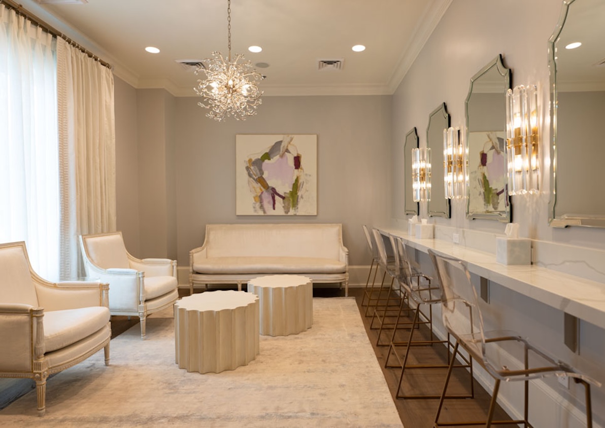 Elegant waiting room with cream armchairs, sofa, round coffee tables, mirrored wall with modern sconces, and transparent chairs along white counter.