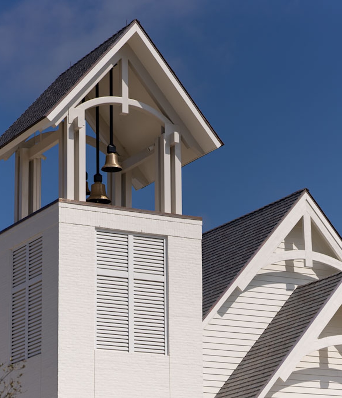 White bell tower with two bronze bells under a steep shingled roof against a clear blue sky.