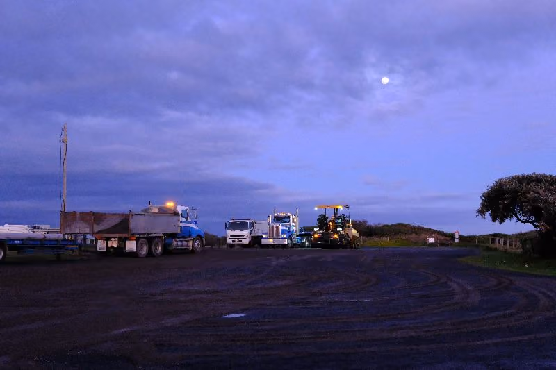 Heavy trucks and road equipment parked under a twilight sky with visible moon, surrounded by open space and silhouetted trees.