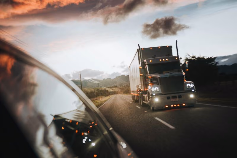 Semi truck driving toward camera on two-lane highway at sunset, headlights on, mountains and dramatic clouds in background.