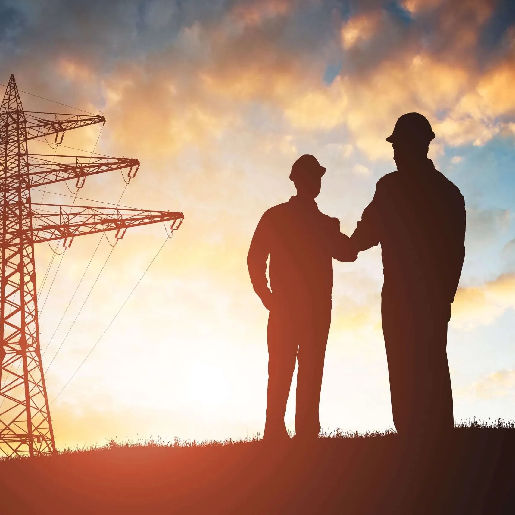 Silhouettes of two workers wearing helmets shaking hands near an electric power transmission tower at sunset.