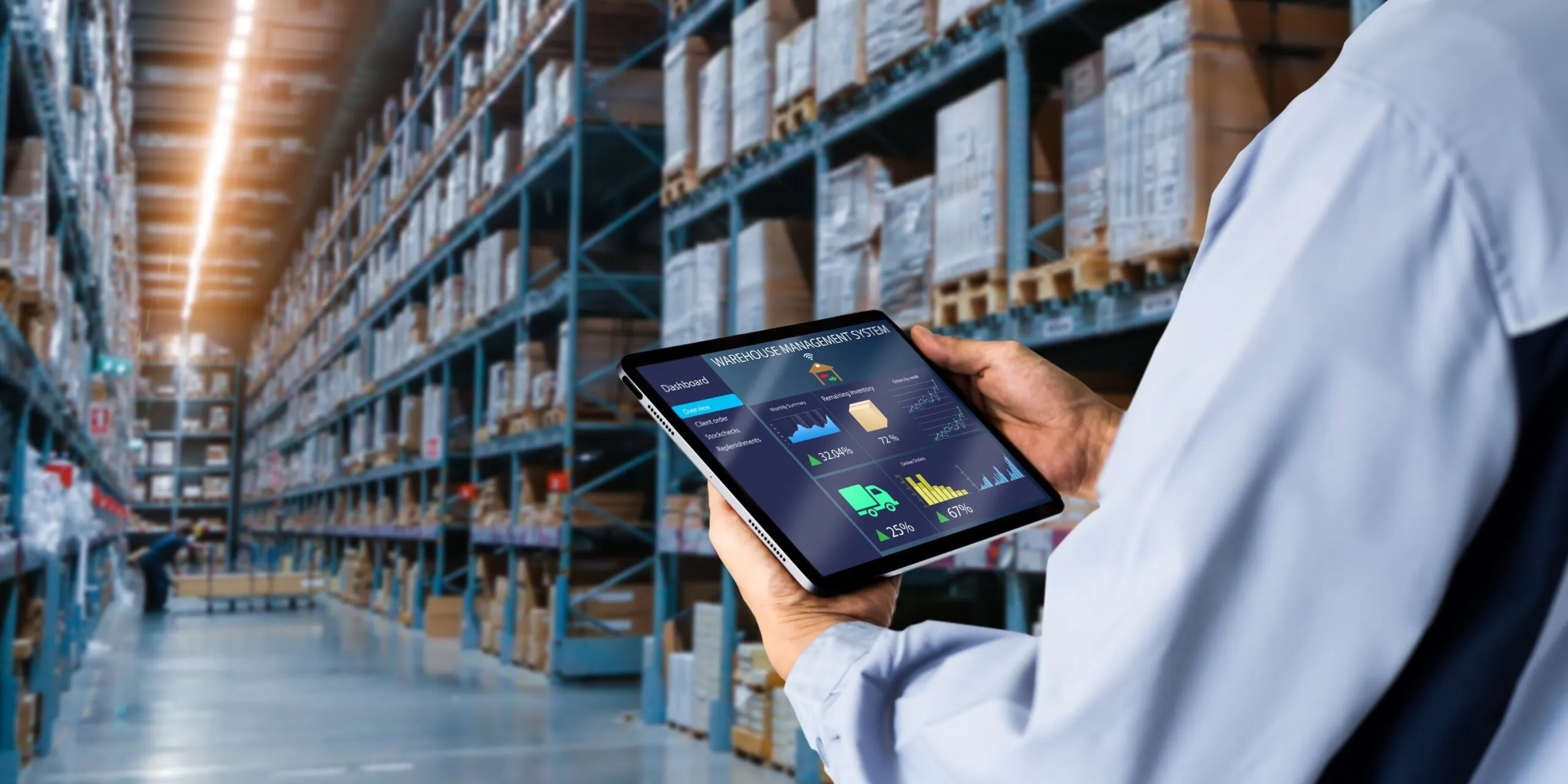 Person using a tablet with a warehouse management system interface in a large warehouse aisle with shelves stacked with boxes.