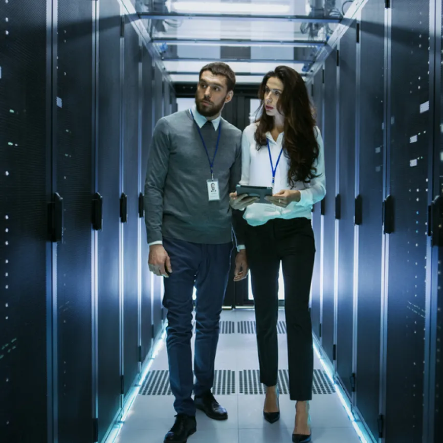 Two IT professionals with ID badges walking through a server room, one holding a tablet.