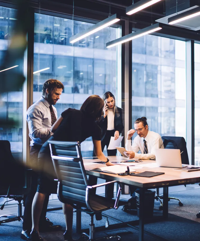 Four business professionals in an office meeting room discussing documents and using a tablet near large windows.