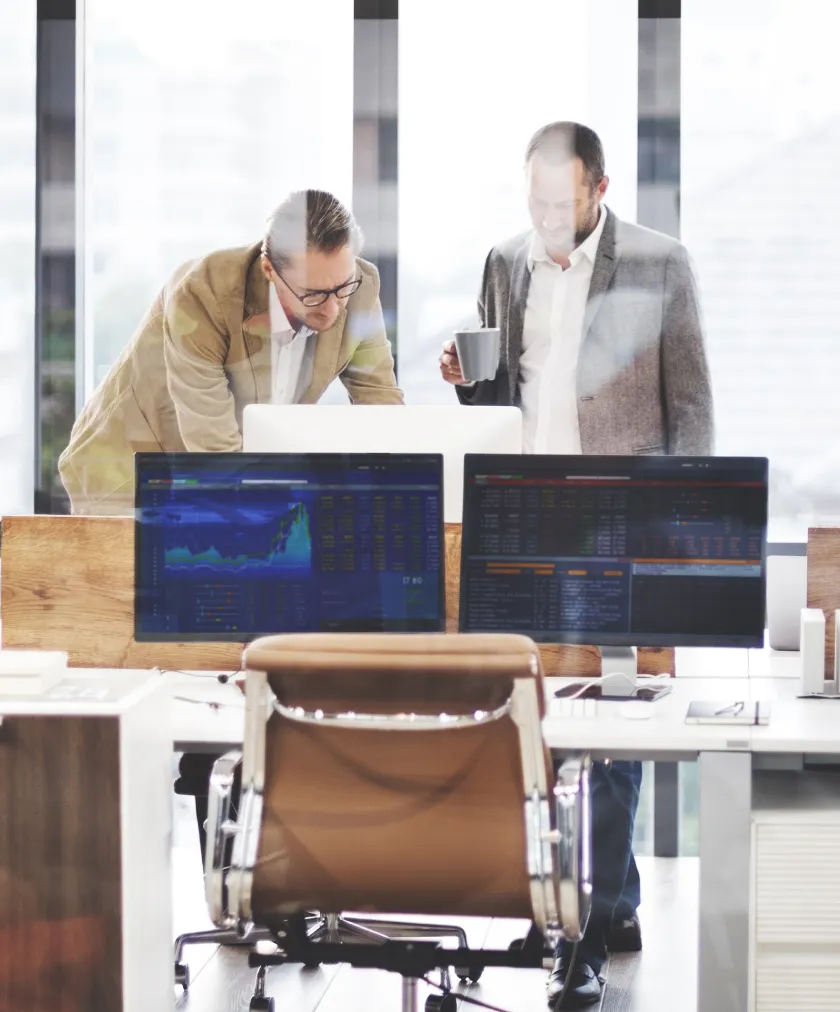 Two business professionals looking at a laptop in a bright office with dual monitors displaying data charts on a desk in the foreground.