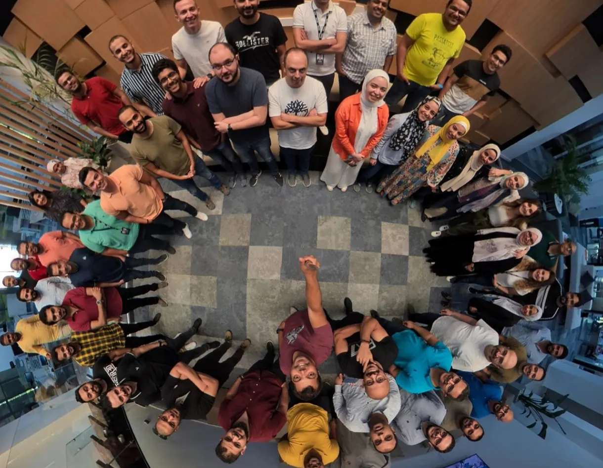 Large group of Incorta team members standing in a circle and smiling up at the camera, captured from an overhead perspective inside a modern office space.