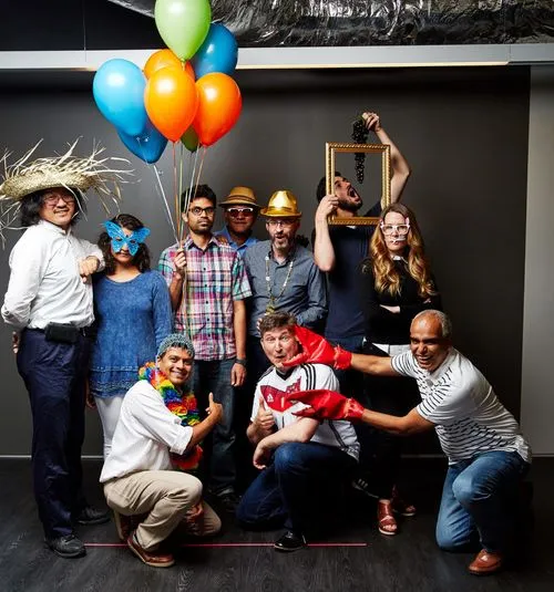 Group of colleagues dressed in fun costumes and props, including hats, masks, balloons, and photo-frame accessories, posing playfully together in an office setting.