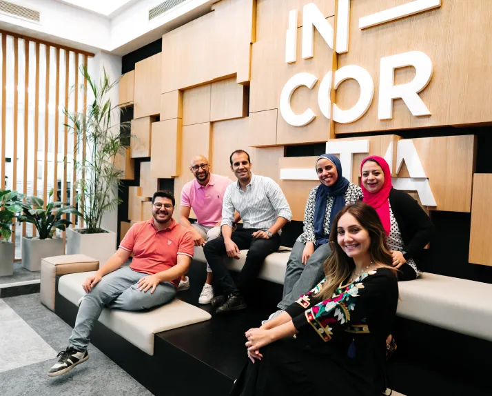 Group of Incorta team members smiling and sitting together in a modern office lounge area with wood panel wall and large Incorta logo in the background.