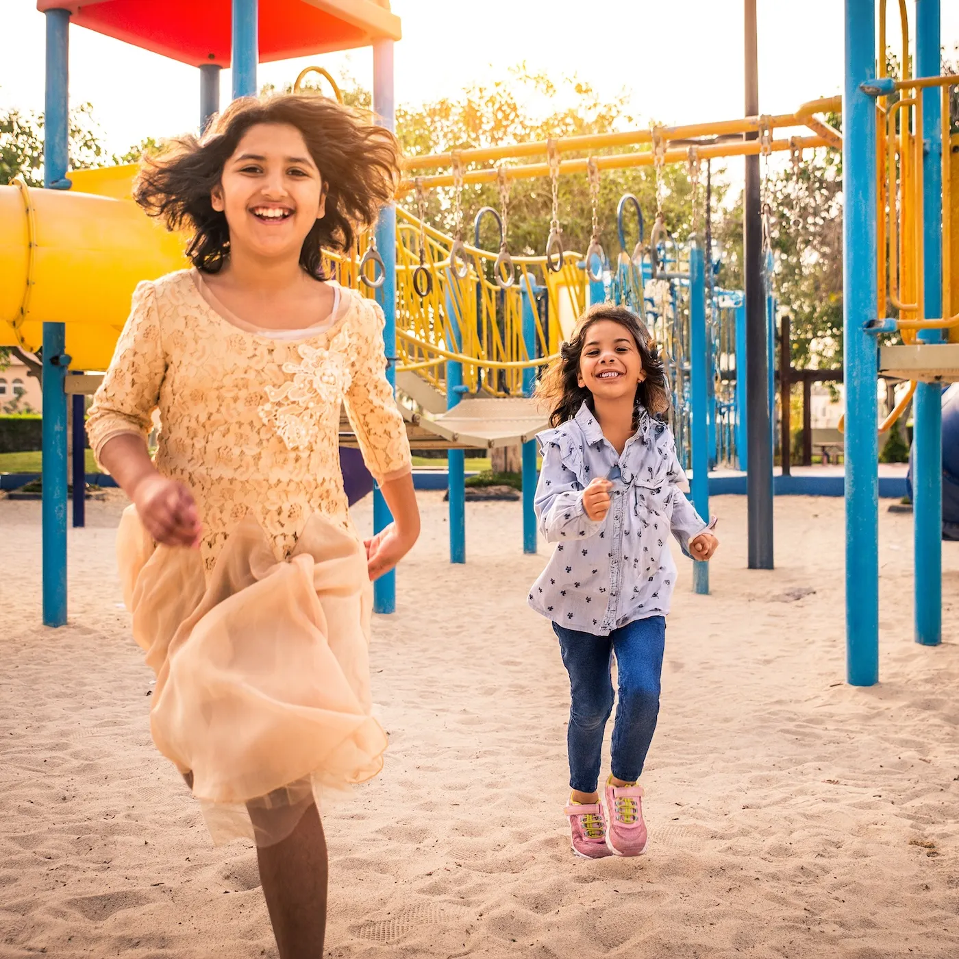 Two young girls laughing and running at a colorful playground, surrounded by slides, climbing structures, and sand underfoot on a sunny day.