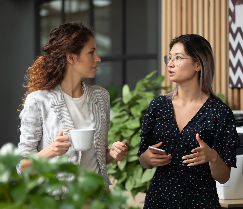 Two professional women having a focused conversation in a modern office with greenery, one holding a coffee mug and the other a smartphone.