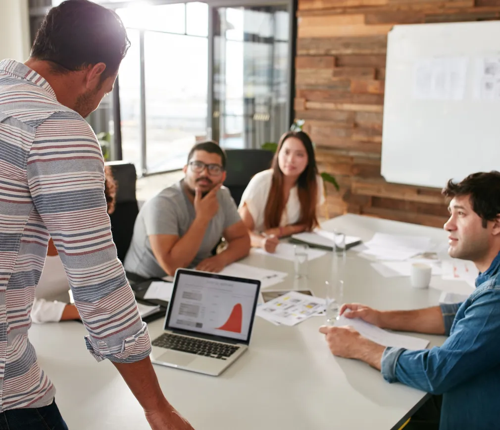 Team gathered in a modern office around a conference table, listening to a presenter with a laptop displaying a graph and analytics data.