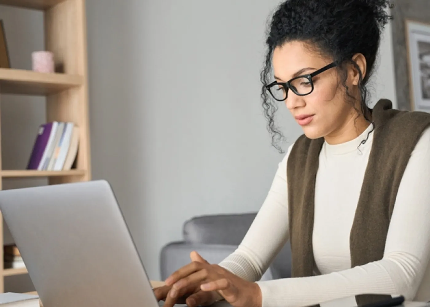 Focused woman with curly hair and glasses working on a laptop at home, seated near a bookshelf.