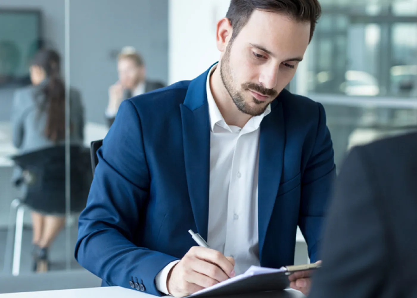 Business professional in a blue suit taking notes during a meeting in a modern office setting.