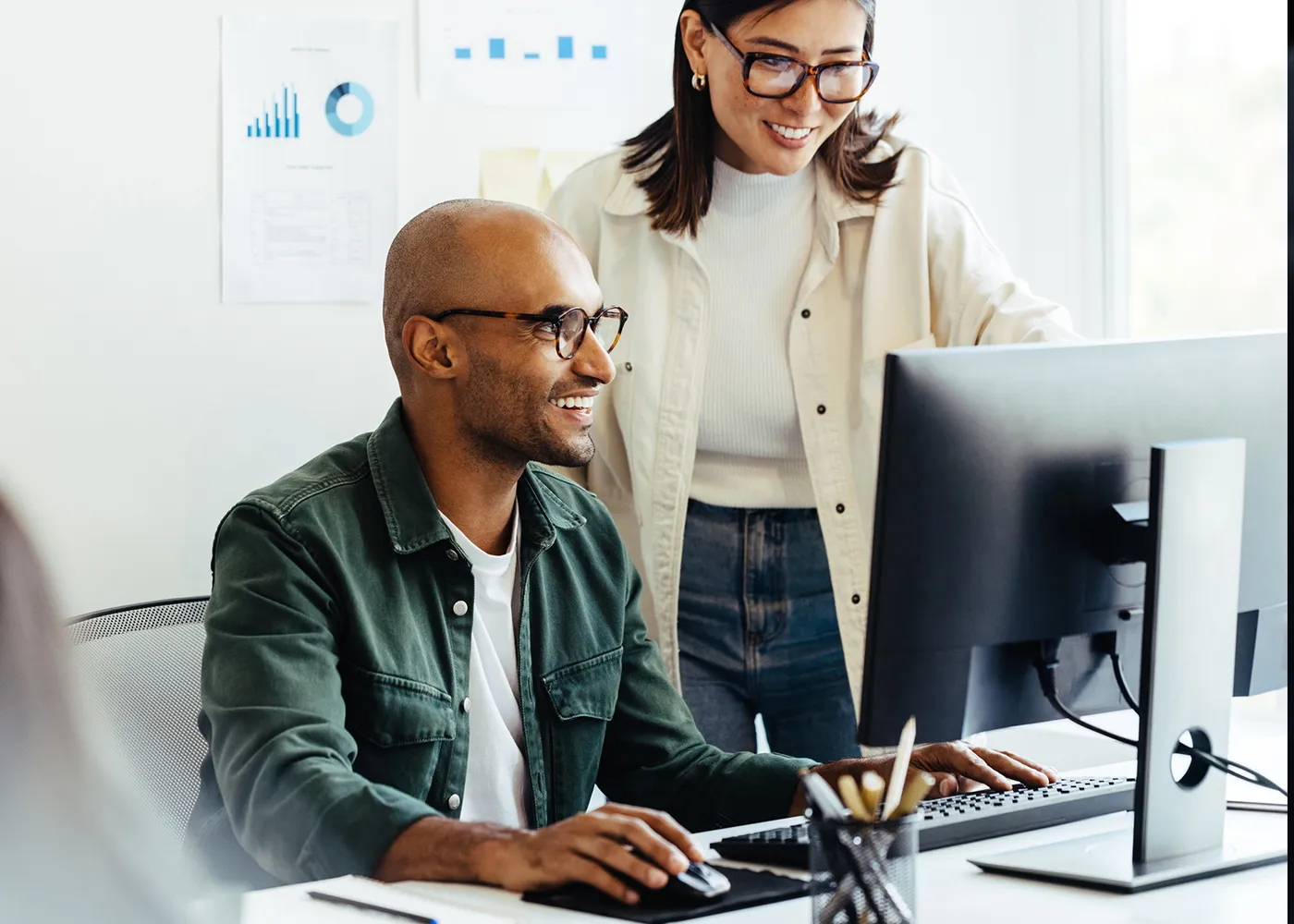 Two coworkers smiling while collaborating at a computer workstation in a bright office, with data charts on the wall in the background, suggesting a productive, analytics-driven environment.