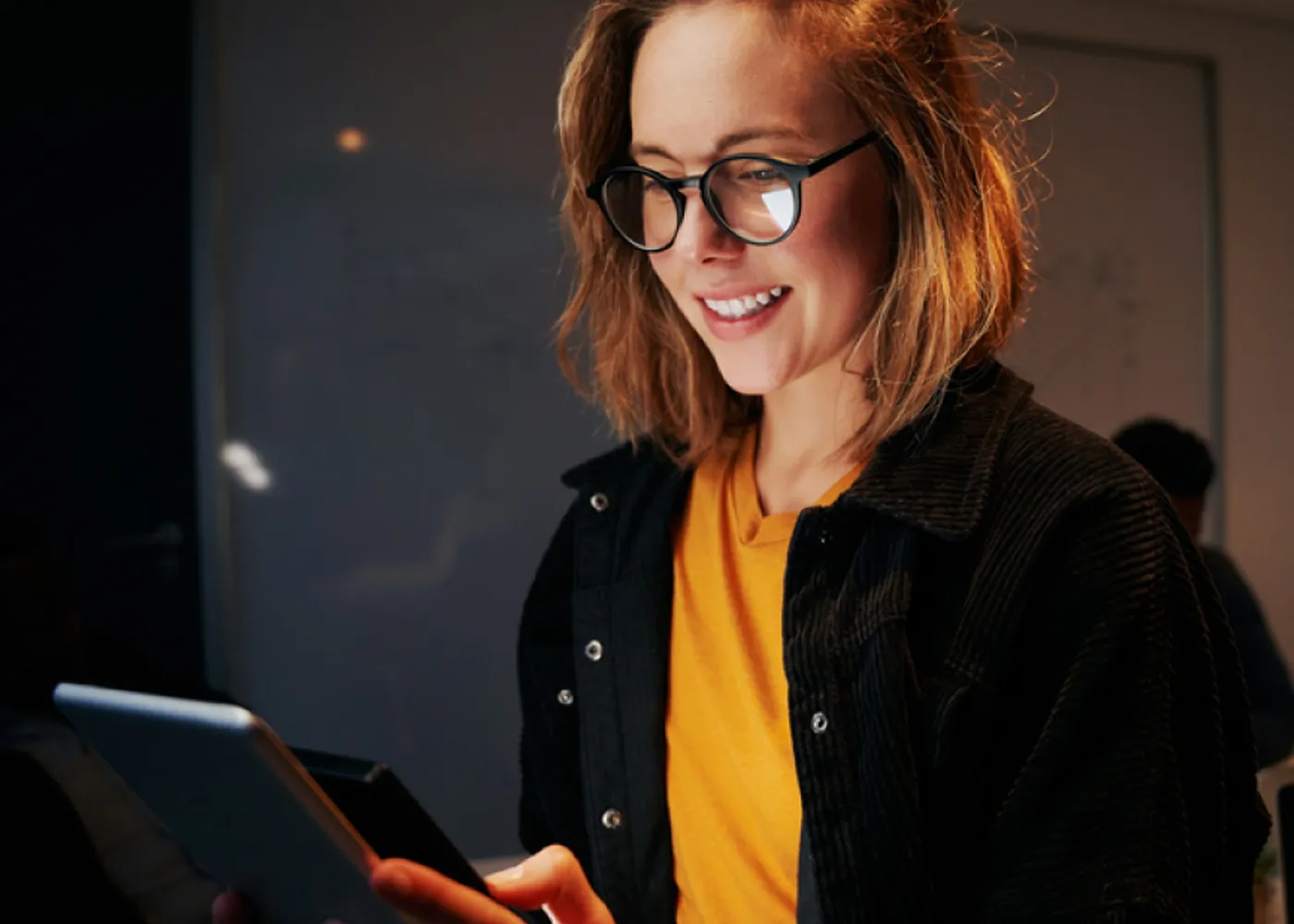 Young woman wearing glasses and a yellow shirt, smiling while using a digital tablet in a dimly lit room, suggesting focus and engagement in a tech or data-driven environment.