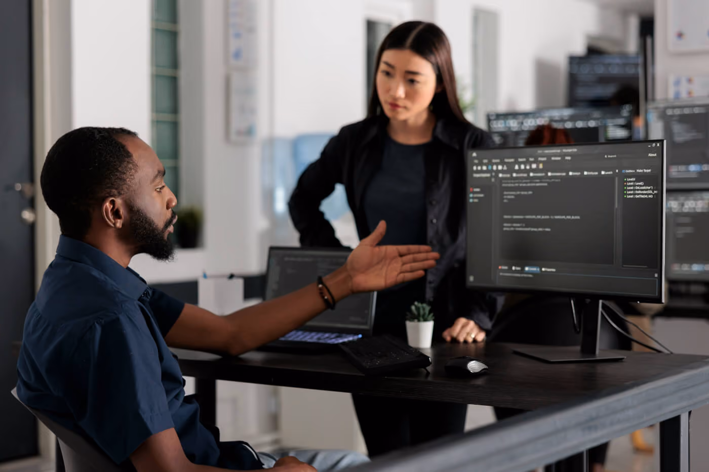 Man seated at desk showing code on a computer monitor to a standing woman in an office.