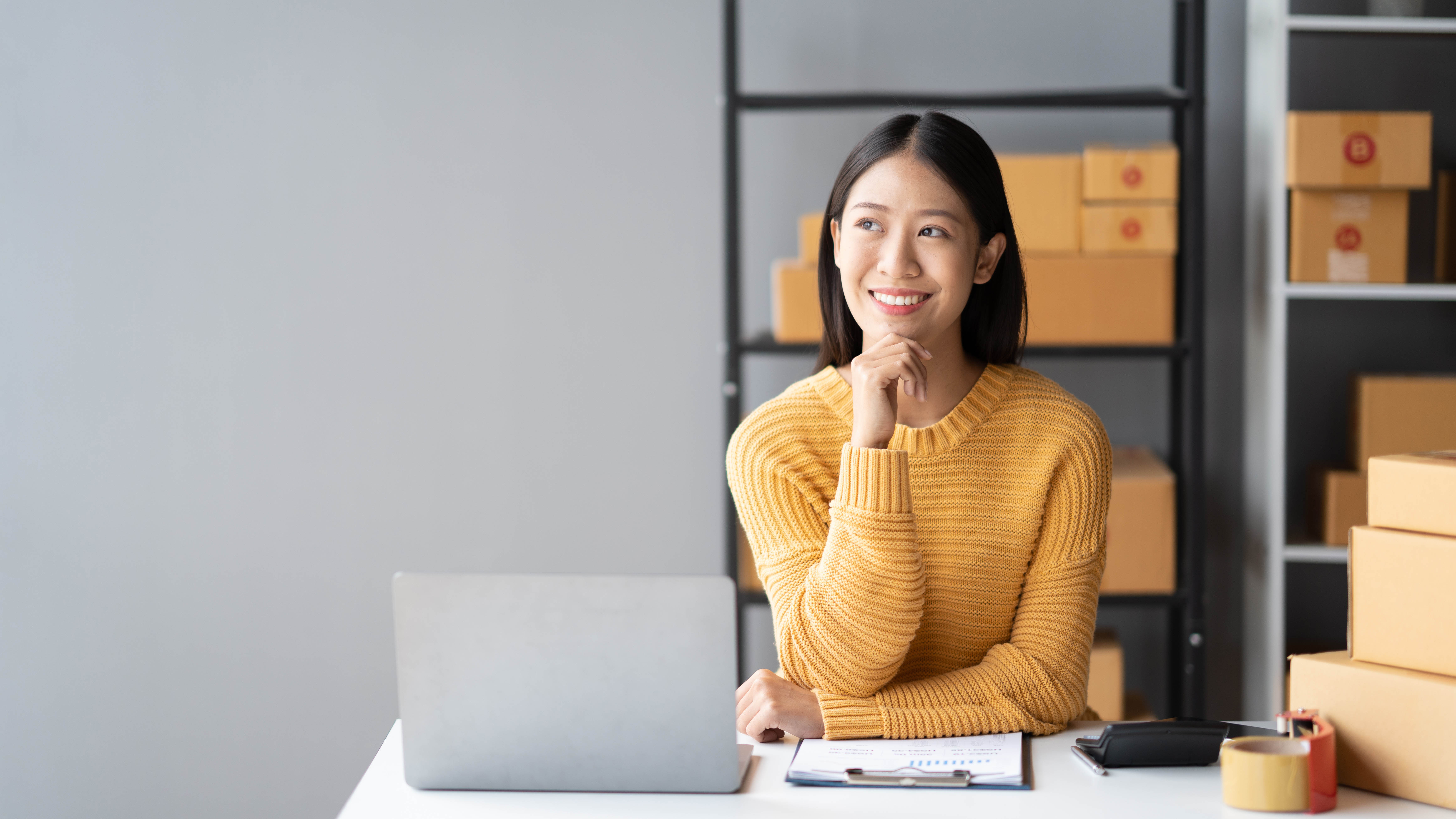 Small business owner at a laptop with shipping boxes behind her, managing her Shopify store after the Summer Editions 2026 updates