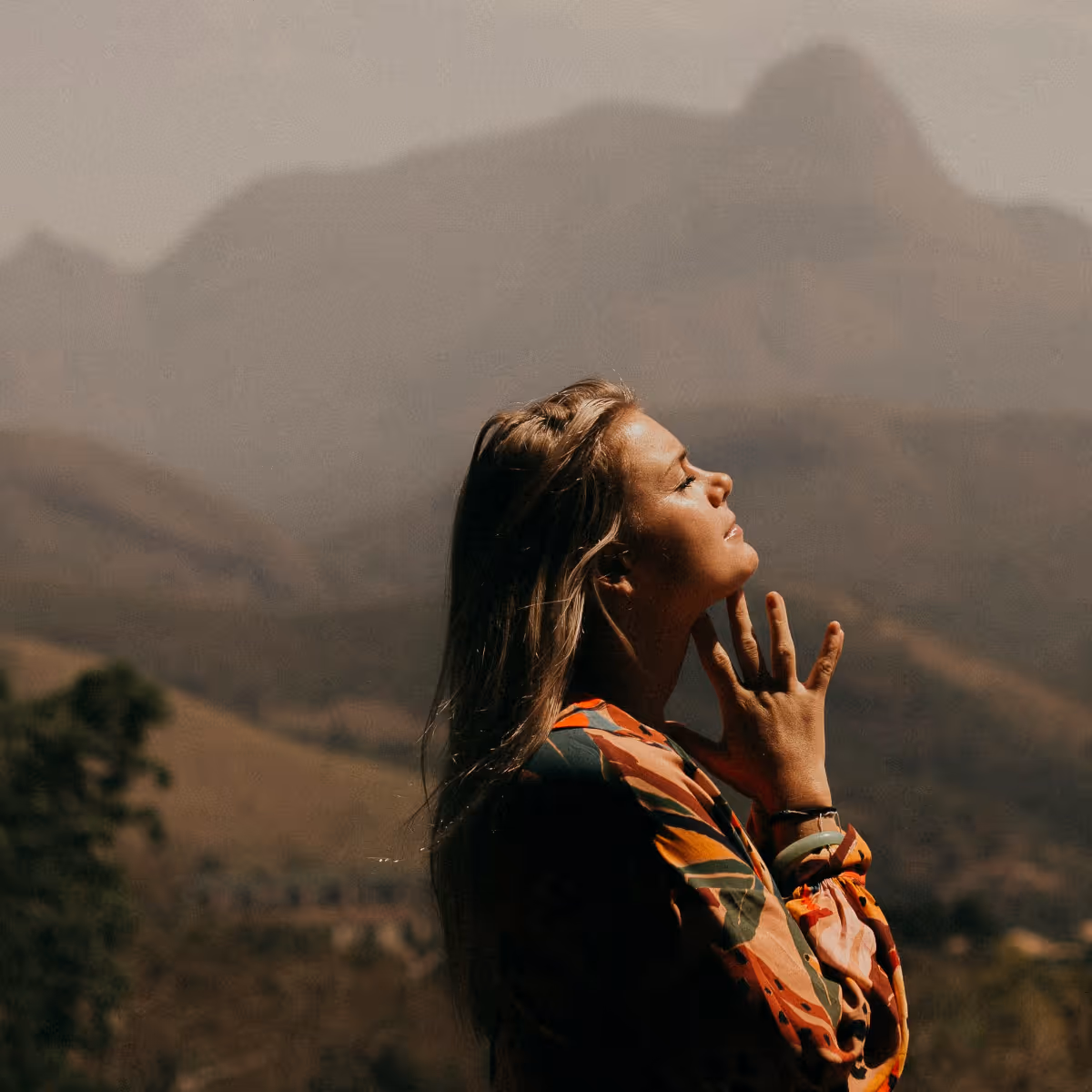 Woman with eyes closed and hands near her neck, standing outdoors with mountains in the background.