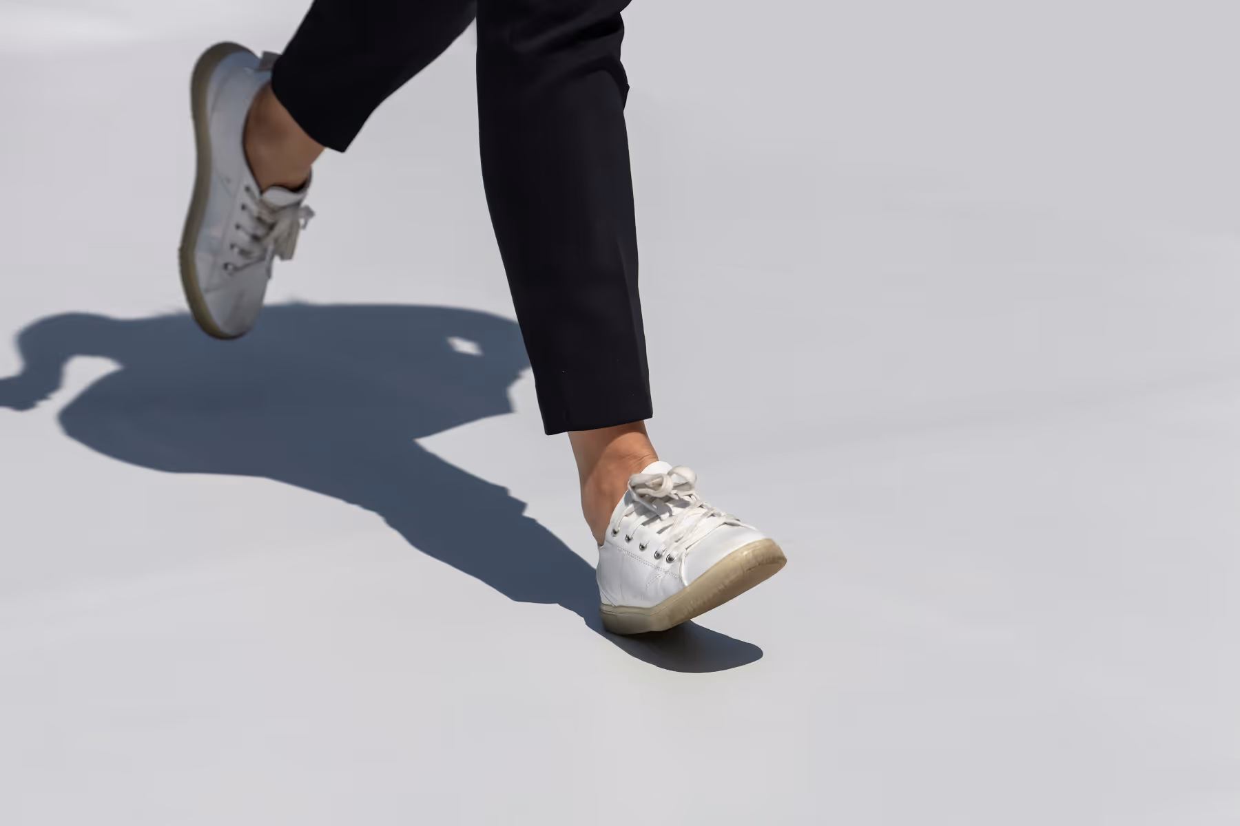 Close-up of feet wearing white sneakers and black pants walking on a light gray surface casting a shadow.