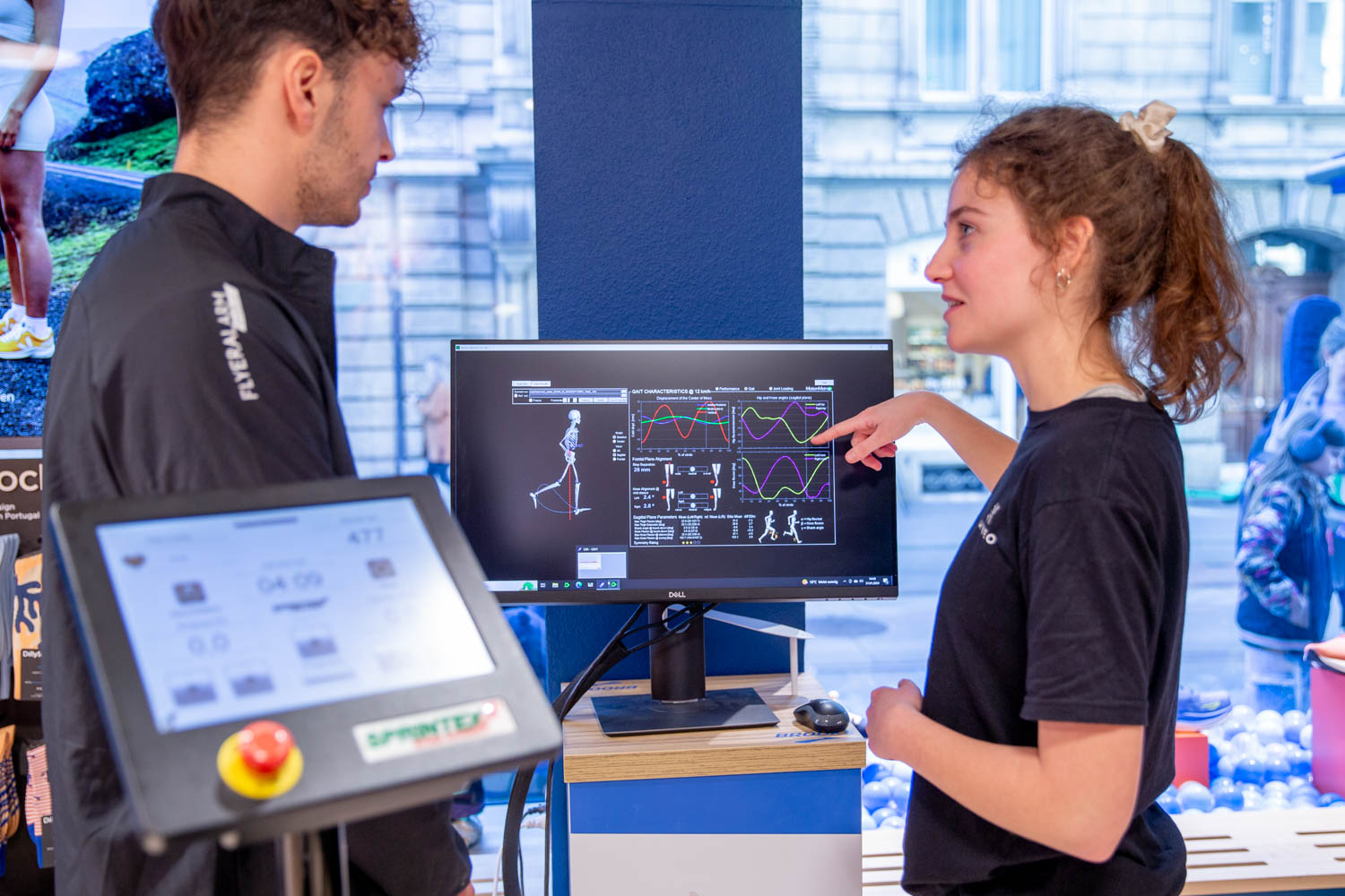 Mitarbeiterin im schwarzen Kineo-Shirt erklärt einem Kunden die Auswertung einer Laufanalyse mit Skelettdarstellung und Kurven am Monitor