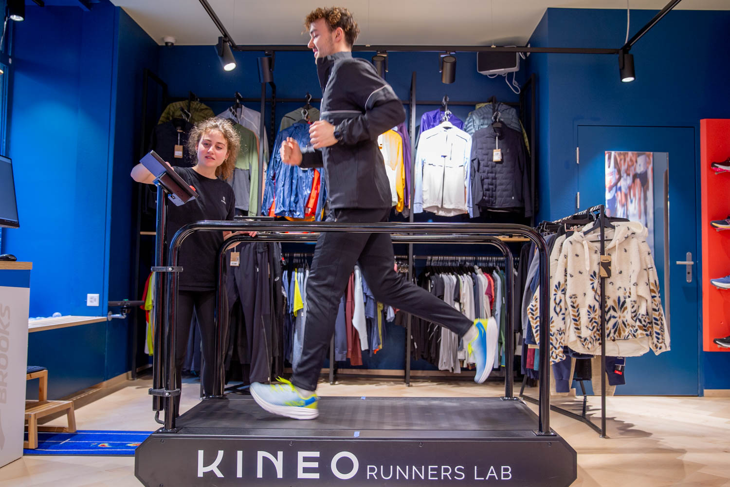 Läufer auf einem Laufband mit dem Schriftzug Kineo Runners Lab, eine Mitarbeiterin filmt den Laufstil mit einem Tablet