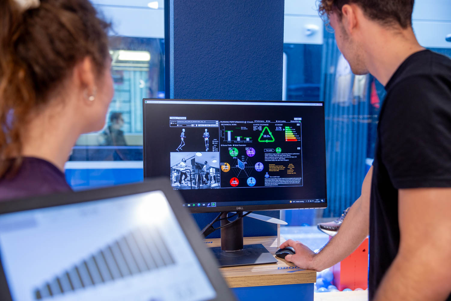 Zwei Personen besprechen vor einem Monitor die Auswertung einer Currex 5D Laufanalyse, im Vordergrund ein Laptop mit Balkendiagramm