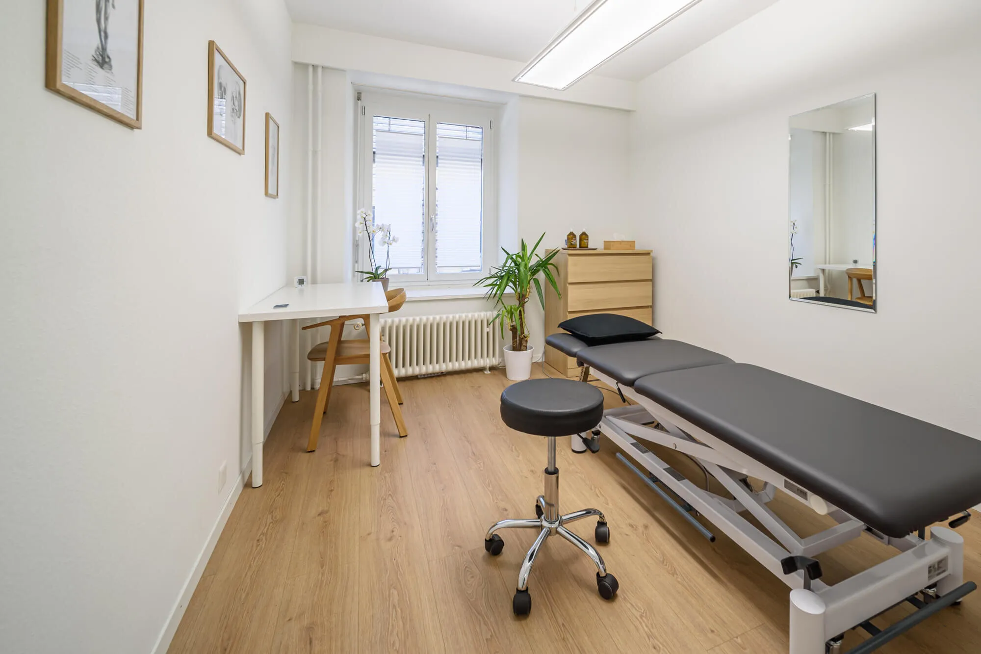 Heller Behandlungsraum bei Kineo Seefeld mit Liege, Hocker, Schreibtisch und Pflanze am Fenster