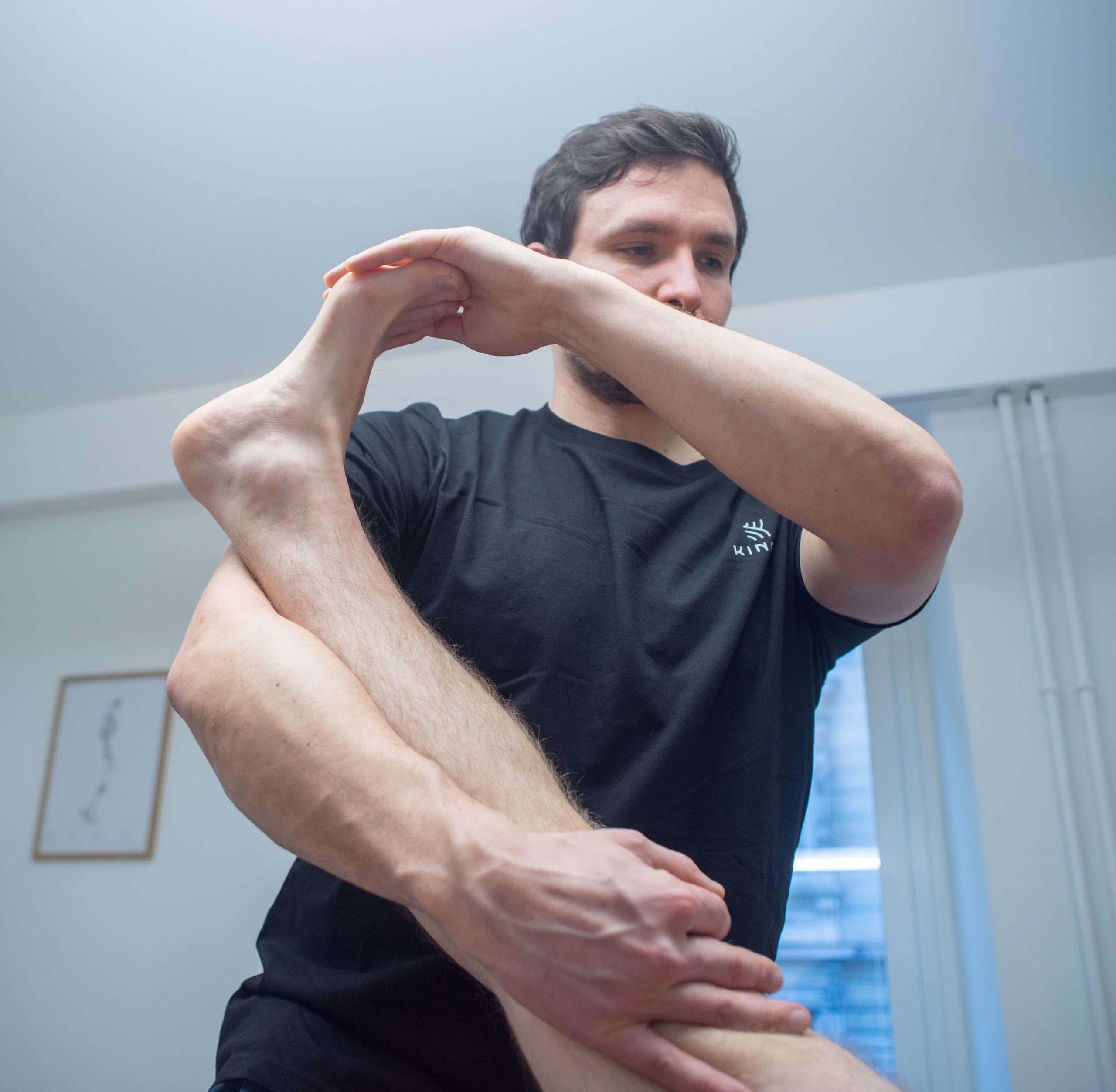 Physiotherapeut im schwarzen Kineo-Shirt dehnt das gestreckte Bein eines liegenden Patienten, der Fuss liegt an seiner Schulter